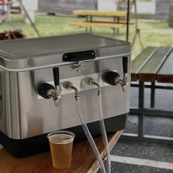 Stainless steel KOMOS jockey box on a wooden picnic table at an outdoor party, surrounded by beer glasses, grilled food, and friends under string lights