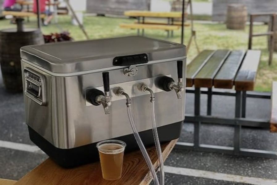 Stainless steel KOMOS jockey box on a wooden picnic table at an outdoor party, surrounded by beer glasses, grilled food, and friends under string lights